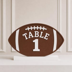 Può includere: Un segnatavolo a forma di palla da football americano marrone con scritte bianche in rilievo che riportano TABLE 1, arricchito da lacci e strisce verticali bianche, montato su una base rettangolare bianca per una posa stabile su una superficie.