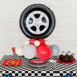 May include: A party table decorated with a racing theme. A large black and white tire-shaped balloon is the centerpiece, surrounded by red, white, and gray balloons. The table has a black and white checkered pattern, with cupcakes, candy, and plates that say "VROOM".