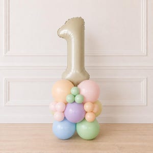 May include: A large beige number one foil balloon standing atop a cluster of smaller latex balloons in pastel shades of peach, pink, lavender, light blue, mint green, and pale yellow, arranged in a decorative column formation for display.