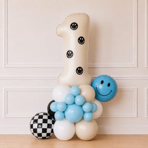 Pu&ograve; includere: Un grande palloncino a forma di numero uno color crema decorato con faccine sorridenti nere, circondato da un gruppo di palloncini bianchi, crema e azzurri, un palloncino rotondo blu con faccina sorridente e un palloncino sferico a scacchi.