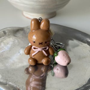 May include: A brown bunny keychain with a pink bow and a strawberry charm. The bunny has a cute, rounded design. The strawberry is pink with green leaves. The keychain is on a silver tray.