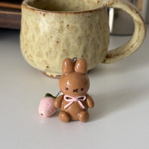May include: A brown bunny charm with a pink bow and a small pink strawberry. The charm is in front of a beige ceramic mug with a large handle. The mug has a speckled texture and a rustic appearance.