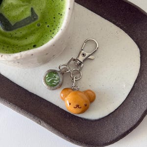 May include: A keychain with a silver clasp and two charms. One charm is a small, round, light green and white design. The other charm is a small, orange bear face. The keychain rests on a white and brown ceramic tray, next to a cup of green matcha.