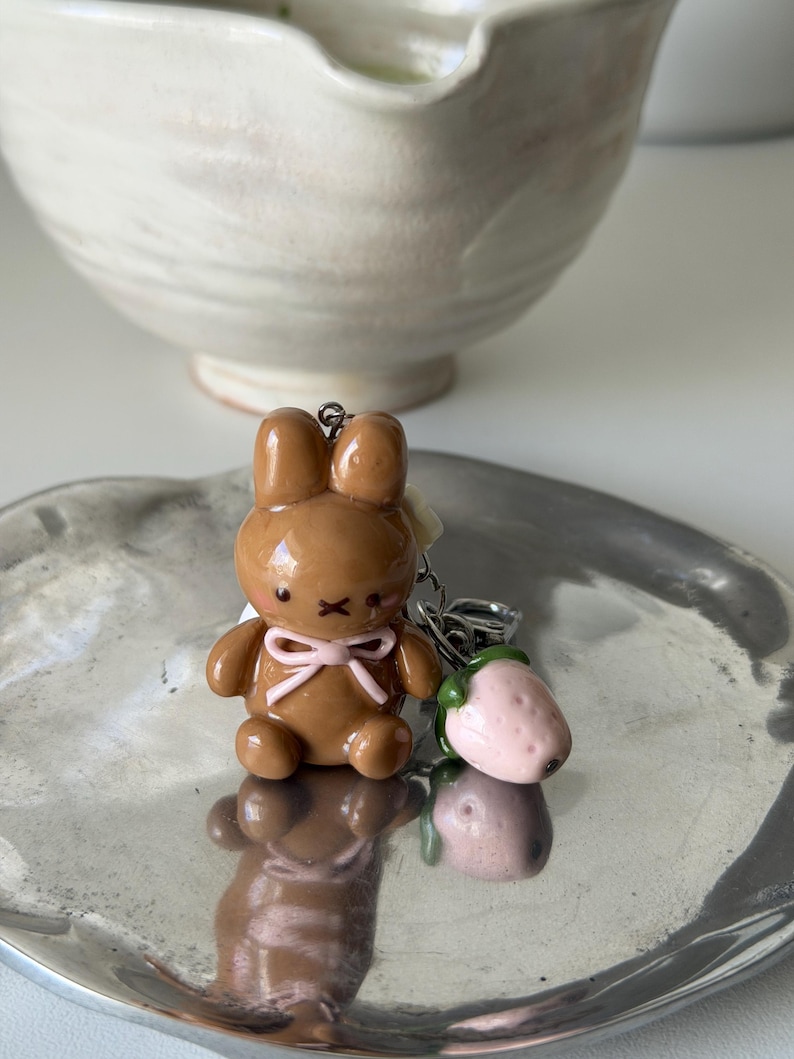 May include: A brown bunny keychain with a pink bow and a strawberry charm. The bunny has a cute, rounded design. The strawberry is pink with green leaves. The keychain is on a silver tray.