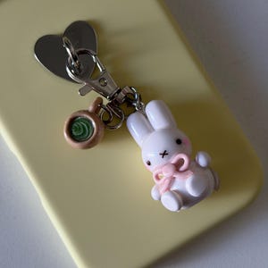 May include: A silver-toned metal keychain featuring a heart-shaped charm, a small brown mug charm with a green swirl center, and a white rabbit figurine charm with pink cheeks, a pink bow, and a black cross-shaped mouth attached to a lobster clasp.