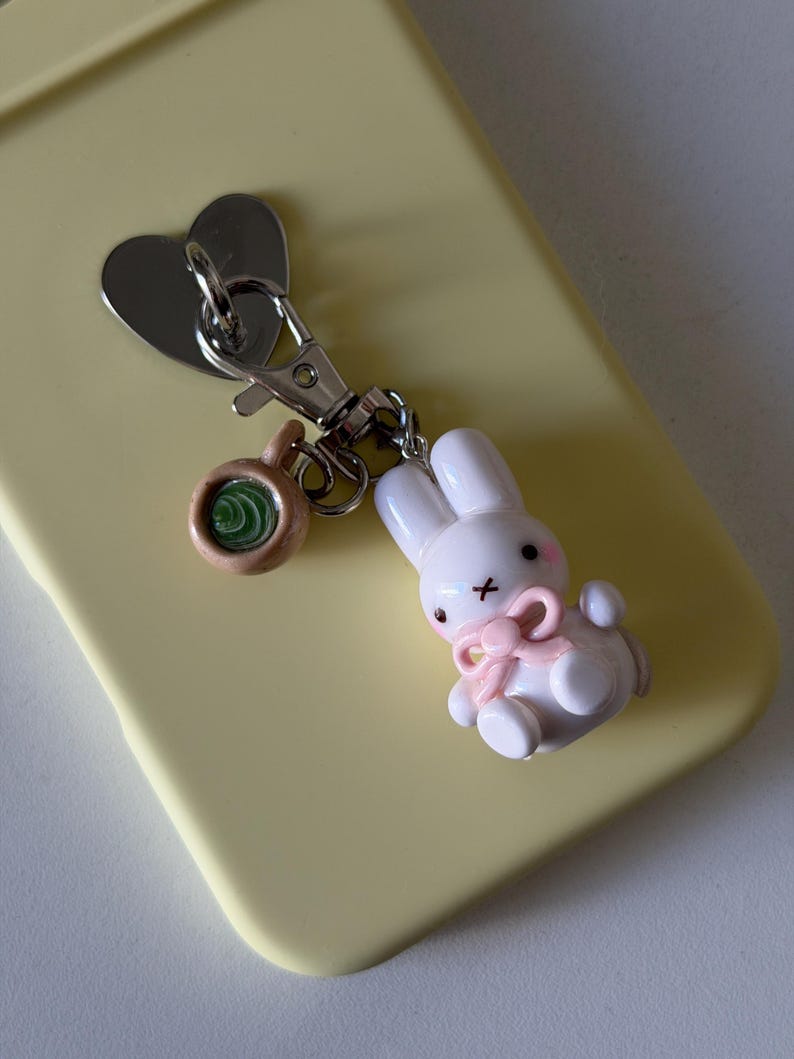May include: A silver-toned metal keychain featuring a heart-shaped charm, a small brown mug charm with a green swirl center, and a white rabbit figurine charm with pink cheeks, a pink bow, and a black cross-shaped mouth attached to a lobster clasp.