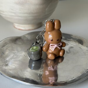 May include: A brown bunny keychain with a pink bow and a small gray sushi keychain. The keychains are on a silver plate. The bunny has a cute expression and is sitting down. The sushi keychain has a green swirl design.