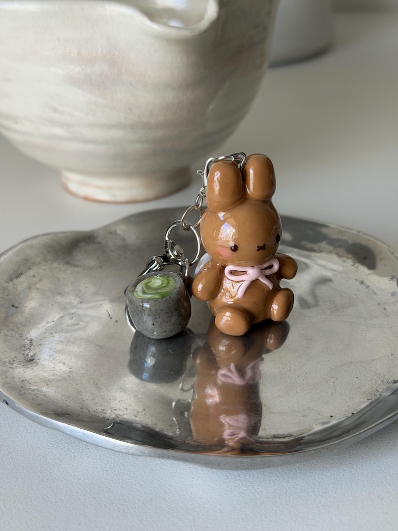 May include: A brown bunny keychain with a pink bow and a small gray sushi keychain. The keychains are on a silver plate. The bunny has a cute expression and is sitting down. The sushi keychain has a green swirl design.