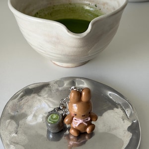 May include: A brown bunny keychain with a pink bow and a small matcha-themed charm sits on a silver plate. A ceramic matcha bowl with green tea is in the background, suggesting a tea-themed product.