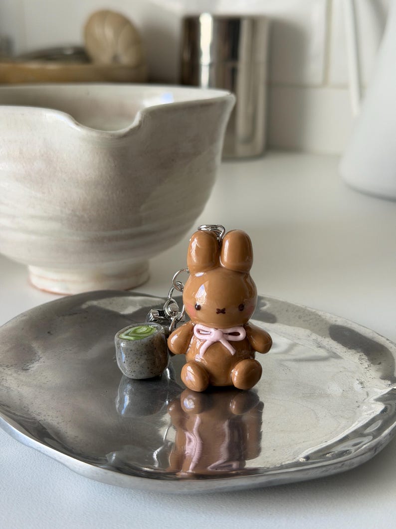 May include: A brown bunny keychain with a pink bow and a small green and gray accessory. The bunny is sitting on a silver tray. The background includes a white ceramic bowl and other kitchen items.