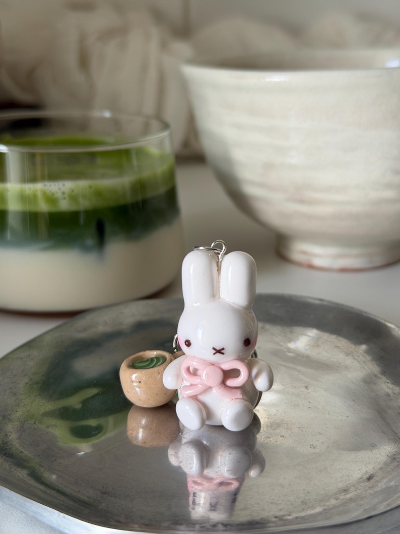 May include: A white rabbit figurine keychain featuring a glossy finish, a light pink bow around its neck, and a small tan-colored cup accessory with green contents attached to the side, all resting on a reflective silver-toned metal surface.