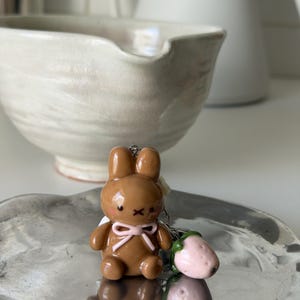 May include: A brown bunny keychain with a pink bow and black details, next to a pink strawberry charm. The bunny is on a silver tray, with a white bowl and pitcher in the background.