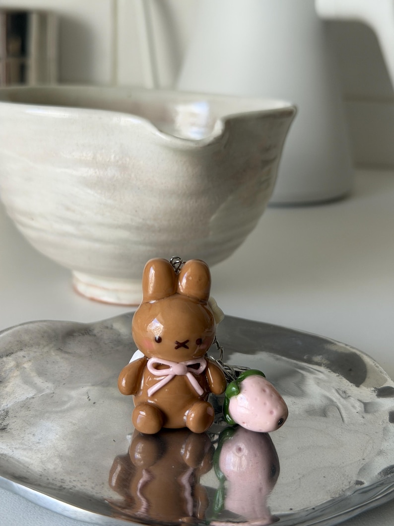 May include: A brown bunny keychain with a pink bow and black details, next to a pink strawberry charm. The bunny is on a silver tray, with a white bowl and pitcher in the background.