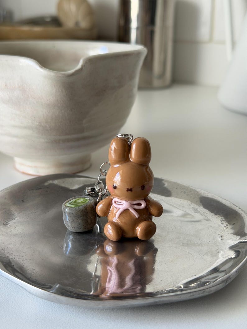 May include: A brown bunny keychain with a pink bow and a small, round charm. The bunny is on a silver-colored tray. A ceramic bowl is in the background.