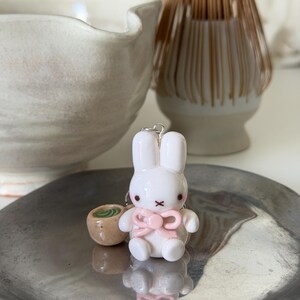 May include: A white rabbit figurine with a pink bow and a small brown cup containing green liquid. The rabbit has black eyes and a black X-shaped mouth. It is attached to a silver metal ring, creating a charm that sits on a metallic surface.