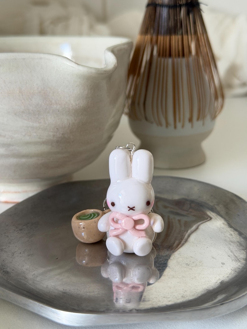 May include: A white rabbit figurine with a pink bow and a small brown cup containing green liquid. The rabbit has black eyes and a black X-shaped mouth. It is attached to a silver metal ring, creating a charm that sits on a metallic surface.