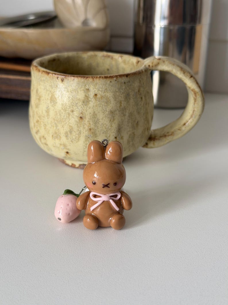 May include: A brown bunny charm with a pink bow and a small pink strawberry. The charm is in front of a beige ceramic mug with a large handle. The mug has a speckled texture and a rustic appearance.