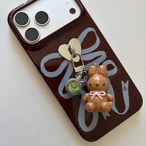 May include: A brown phone case with a light blue ribbon design and a small brown bunny charm with a pink bow. A silver heart-shaped charm and a green and white charm are also attached.