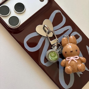 May include: A brown phone case with a light blue ribbon design and a silver heart-shaped charm. A small brown bunny charm with a pink bow and a green and gray charm are attached to the case.