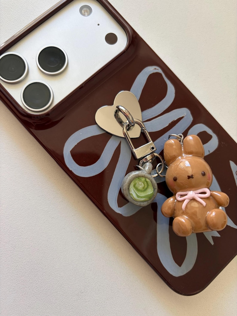 May include: A brown phone case with a light blue ribbon design and a silver heart-shaped charm. A small brown bunny charm with a pink bow and a green and gray charm are attached to the case.