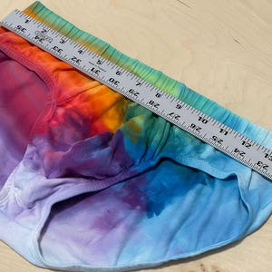 May include: Tie-dye underwear in a rainbow of colors, including red, orange, yellow, green, blue, and purple. A metal ruler is placed across the top of the underwear, measuring approximately 14 inches across.