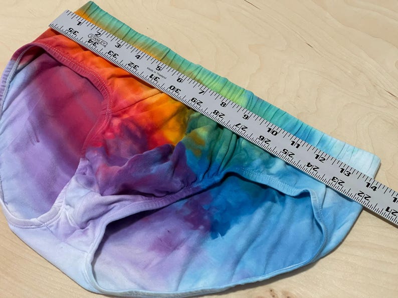 May include: Tie-dye underwear in a rainbow of colors, including red, orange, yellow, green, blue, and purple. A metal ruler is placed across the top of the underwear, measuring approximately 14 inches across.