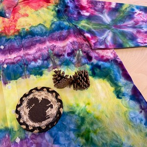 May include: A tie-dye button-down shirt with a colorful swirl pattern in shades of red, yellow, green, blue, and purple. The shirt has a pocket on the left side and a brown woven coaster with a squirrel design in the center.
