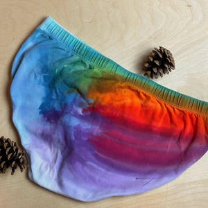 May include: A pair of underwear with a vibrant tie-dye pattern in shades of blue, green, orange, and purple. The elastic waistband is green. Two pinecones are placed near the underwear on a light wooden surface.
