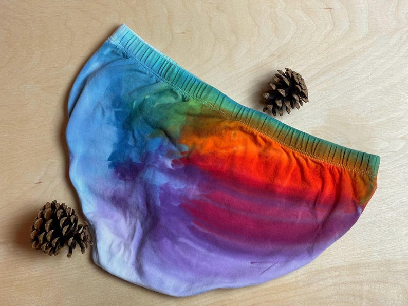 May include: A pair of underwear with a vibrant tie-dye pattern in shades of blue, green, orange, and purple. The elastic waistband is green. Two pinecones are placed near the underwear on a light wooden surface.