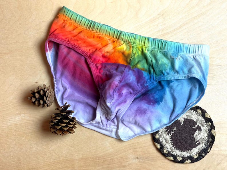 May include: Rainbow tie-dye briefs with a blue elastic waistband. The underwear features a vibrant blend of colors, including red, orange, yellow, green, and blue. The briefs are displayed on a wooden surface with pine cones and a coaster.