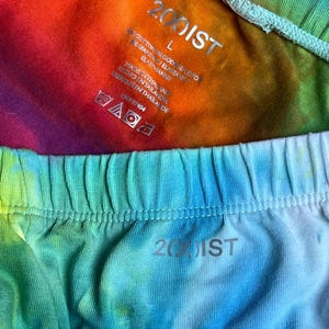 May include: Close-up of two pairs of tie-dye underwear. One pair is a vibrant mix of rainbow colors, and the other is a blend of blue and green hues. The brand name "2(00)IST" is visible on both pairs.
