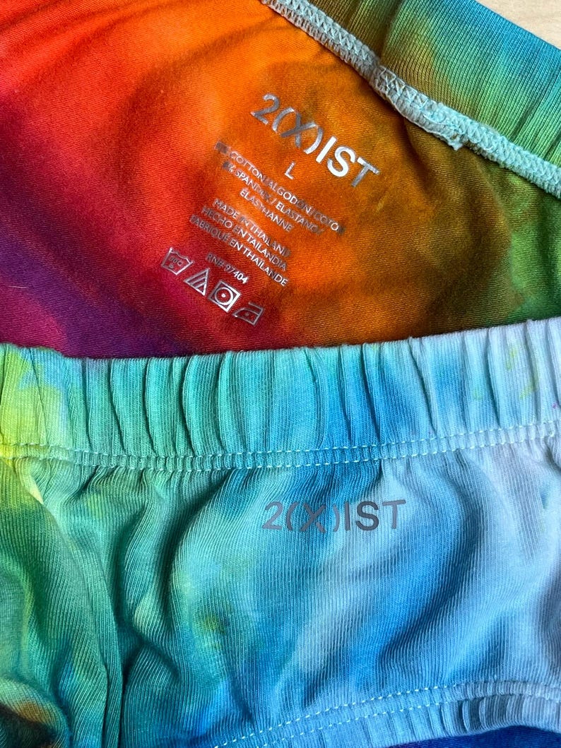 May include: Close-up of two pairs of tie-dye underwear. One pair is a vibrant mix of rainbow colors, and the other is a blend of blue and green hues. The brand name "2(00)IST" is visible on both pairs.