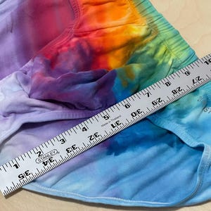 May include: Tie-dye shorts in a rainbow of colors, including purple, blue, orange, and green. A ruler shows a measurement of approximately 35 inches. The shorts have an elastic waistband and are made of a soft fabric.