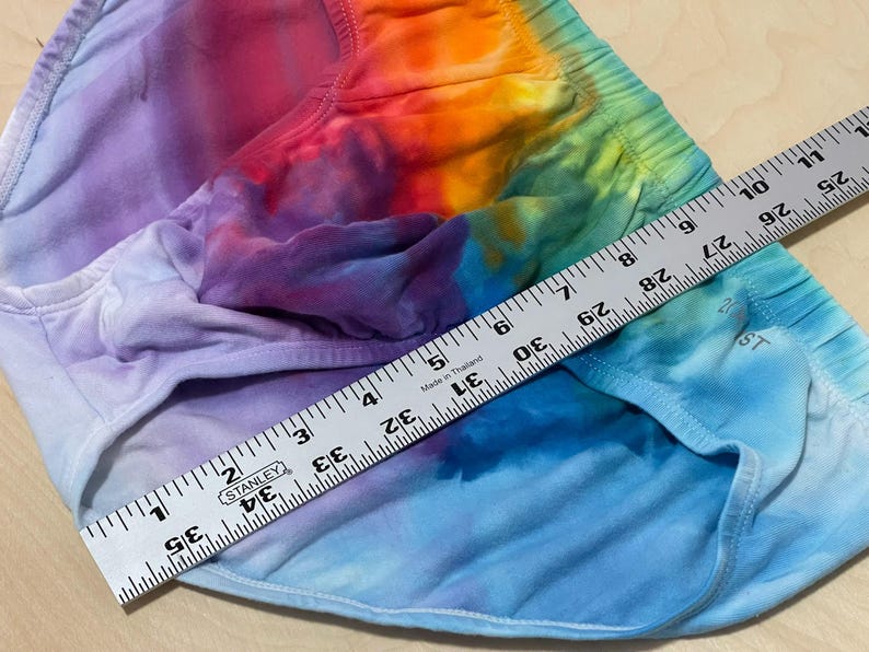 May include: Tie-dye shorts in a rainbow of colors, including purple, blue, orange, and green. A ruler shows a measurement of approximately 35 inches. The shorts have an elastic waistband and are made of a soft fabric.