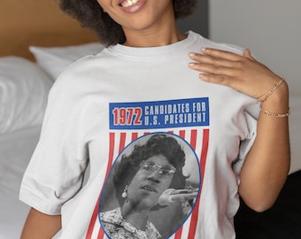 May include: A light gray t-shirt featuring a graphic of Shirley Chisholm, a candidate for U.S. President in 1972. The design includes the text "1972 CANDIDATES FOR U.S. PRESIDENT" and "SHIRLEY CHISHOLM" in red, white, and blue.