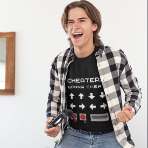 May include: A person wearing a black t-shirt with the text "Cheater's Gonna Cheat" and a pixelated image of a video game controller. The person is holding a black video game controller in their left hand and is smiling and looking at the camera.