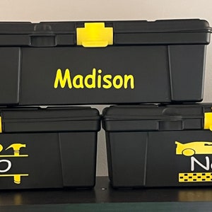 May include: Three black plastic storage bins with yellow lids and personalized labels. The top bin says "Madison", the middle bin says "Teo" with a row of tools, and the bottom bin says "Nash" with a yellow race car.