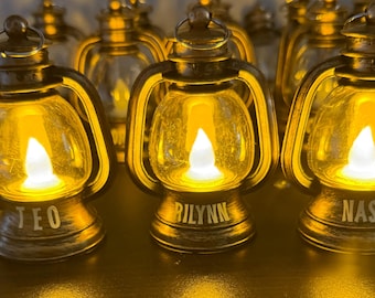 May include: A collection of gold-toned lanterns with a warm, glowing LED flame. Each lantern has a clear glass panel and a handle. The bases are inscribed with names such as "TEO", "RILYNN", and "NASH".