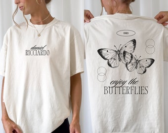 May include: A white t-shirt with the text "daniel ricciardo" on the front and "enjoy the butterflies" with a butterfly graphic on the back. The shirt has a relaxed fit and short sleeves.