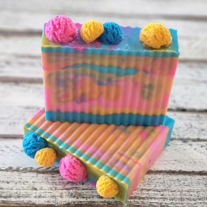 May include: Two bars of handmade soap with a colorful swirl design. The soap is topped with small, colorful balls.
