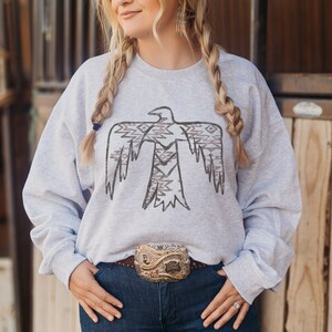 May include: A gray sweatshirt with a stylized black and brown eagle design. The eagle has a geometric pattern on its wings. The sweatshirt is worn by a person with long blonde braids. The person is wearing a belt with a large silver buckle.