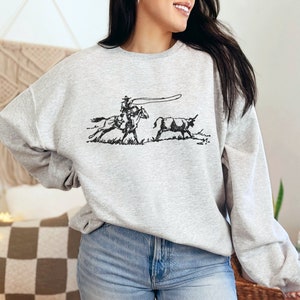 Peut inclure: Un sweat-shirt gris avec une illustration en noir et blanc d'un cow-boy &agrave; cheval qui l&acirc;che un lasso sur un taureau.