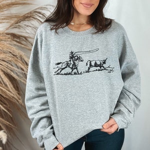 Peut inclure: Un sweat-shirt gris avec une illustration en noir et blanc d'un cow-boy &agrave; cheval qui l&acirc;che un lasso sur un taureau.