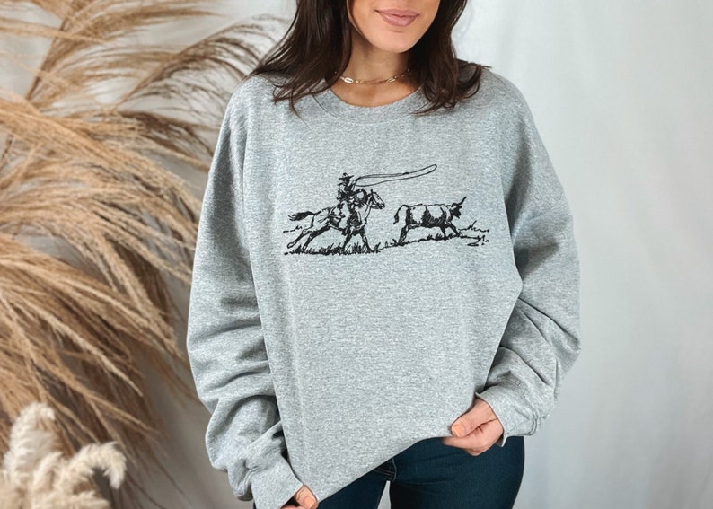 Peut inclure: Un sweat-shirt gris avec une illustration en noir et blanc d'un cow-boy &agrave; cheval qui l&acirc;che un lasso sur un taureau.