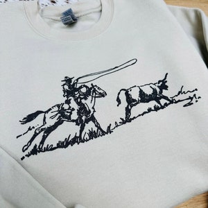 Peut inclure: Un sweat-shirt beige clair avec une illustration en noir et blanc d'un cow-boy &agrave; cheval en train de lassoer un taureau.