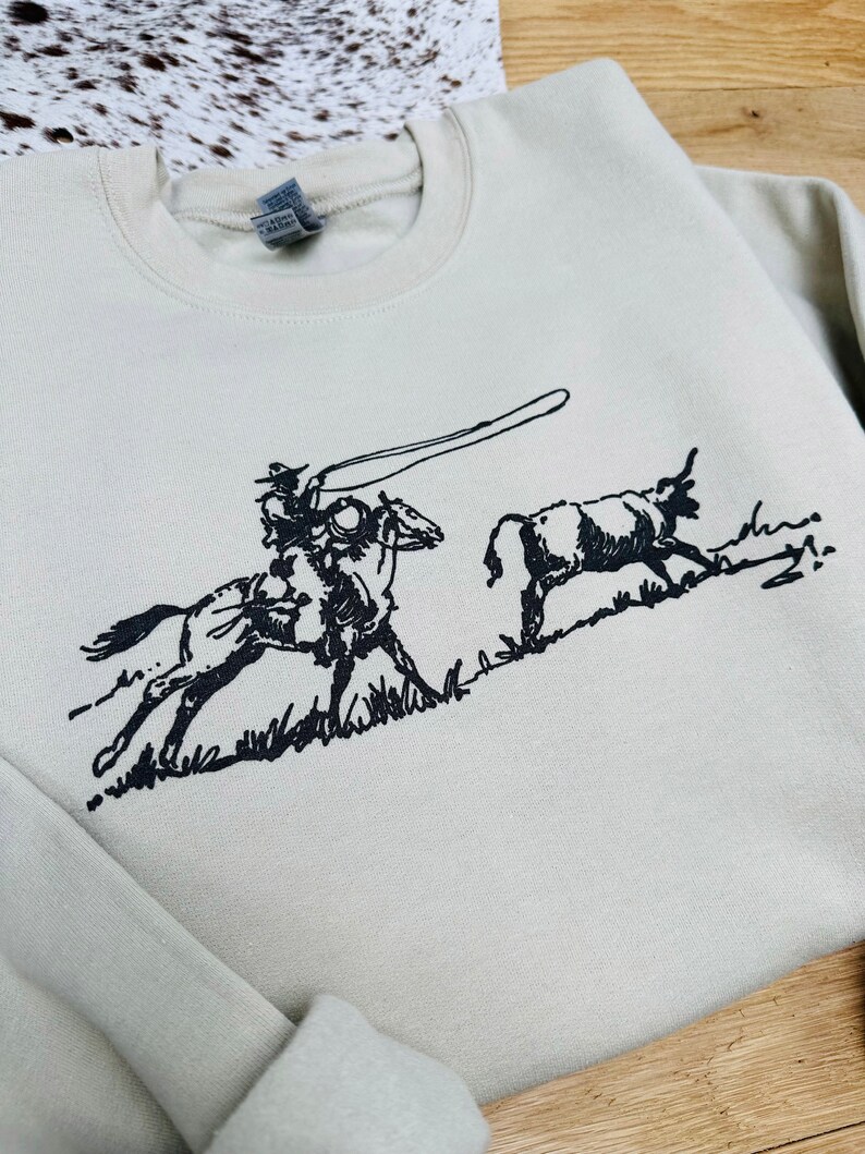 Peut inclure: Un sweat-shirt beige clair avec une illustration en noir et blanc d'un cow-boy &agrave; cheval en train de lassoer un taureau.