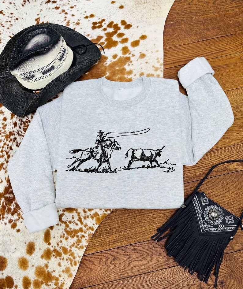 Peut inclure: Un sweat-shirt gris avec une illustration en noir et blanc d'un cow-boy &agrave; cheval qui l&acirc;che un lasso sur un taureau. Le sweat-shirt est pos&eacute; sur un tapis en peau de vache avec un chapeau de cow-boy noir et un sac &agrave; main noir &agrave; franges.