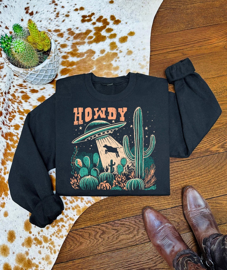 May include: Black sweatshirt with a graphic print of a UFO hovering over a cow in a desert landscape. The text "HOWDY" is printed above the UFO.