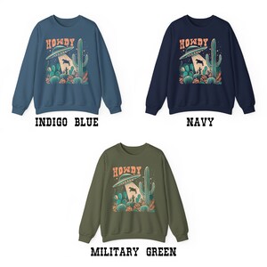May include: Three sweatshirts in different colors: indigo blue, navy blue, and military green. Each sweatshirt features a graphic of a UFO with the word "Howdy" above it, and a cactus in the background.