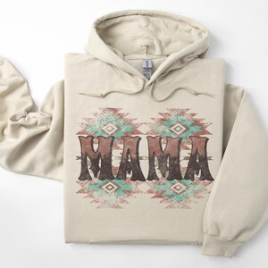 Könnte beinhalten: Beigefarbener Kapuzenpullover mit dem Wort "MAMA" in verblichener brauner Schrift, mittig platziert. Das Wort ist auf einem türkisfarbenen und roten geometrischen Muster angeordnet. Der Hoodie hat eine Kapuze mit Kordelzug und lange Ärmel.